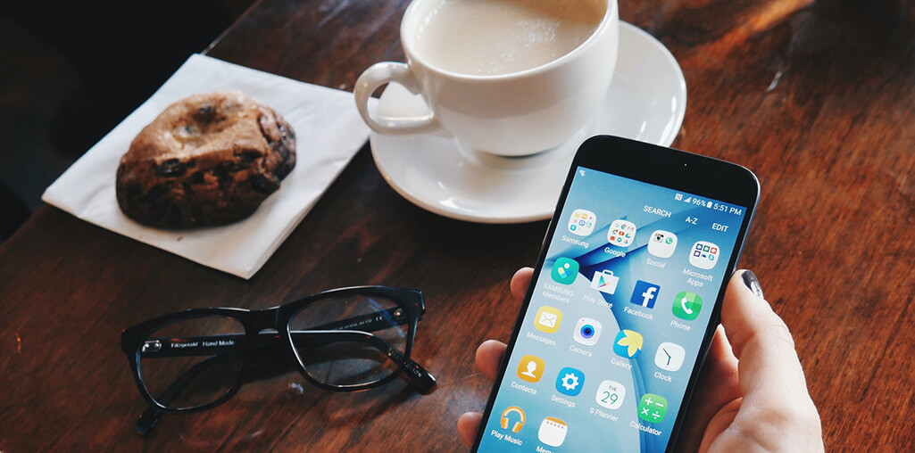 Hand holding an iPhone on a wood table, with coffee and pastry in the background